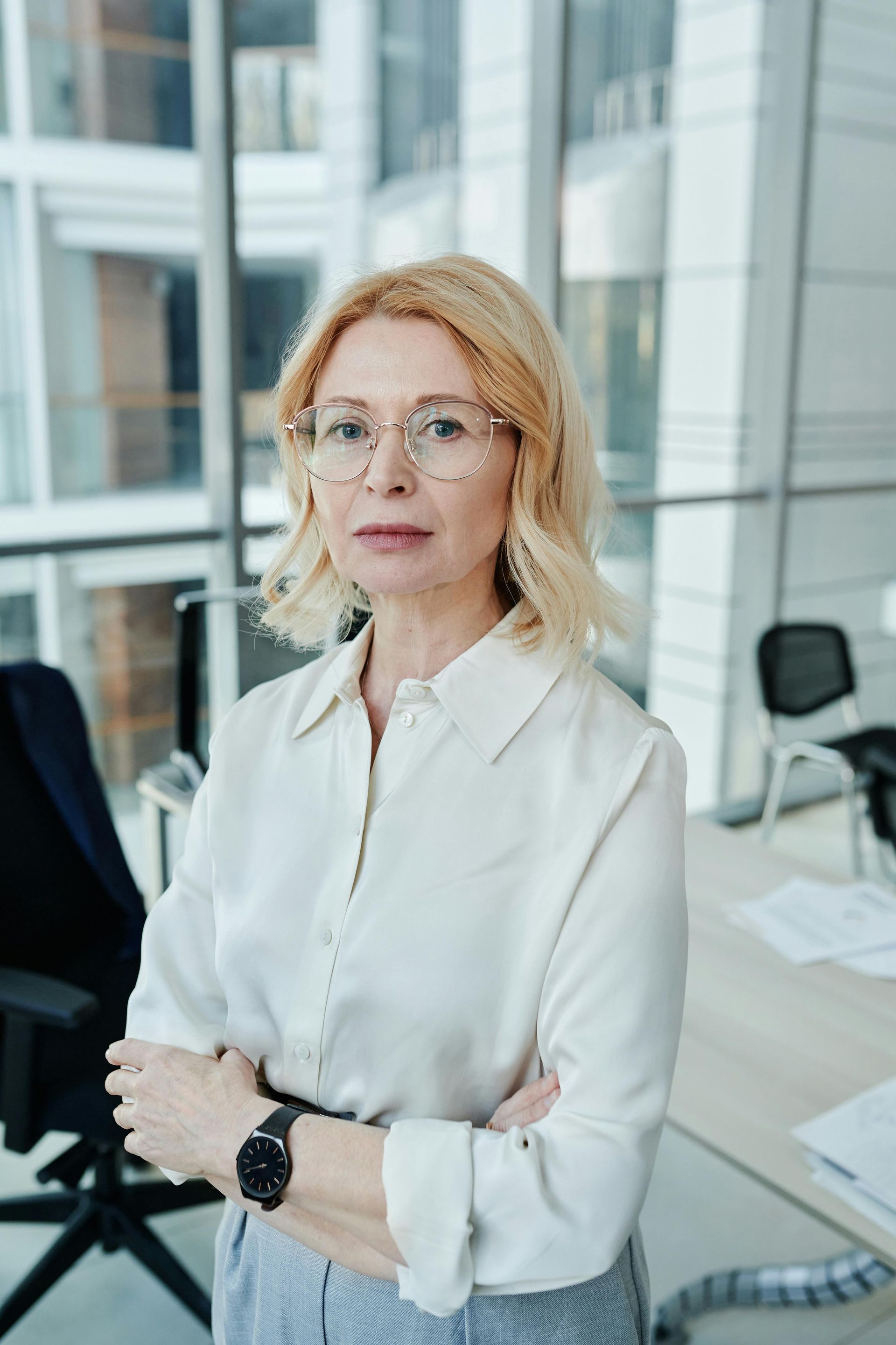 A woman in a white shirt and glasses is standing in an office with her arms crossed.