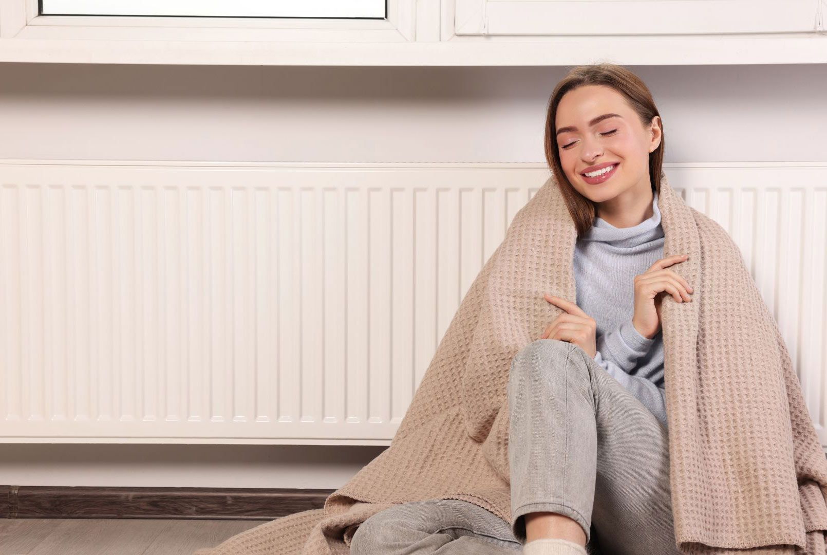 A woman is sitting on the floor wrapped in a blanket in front of a radiator.