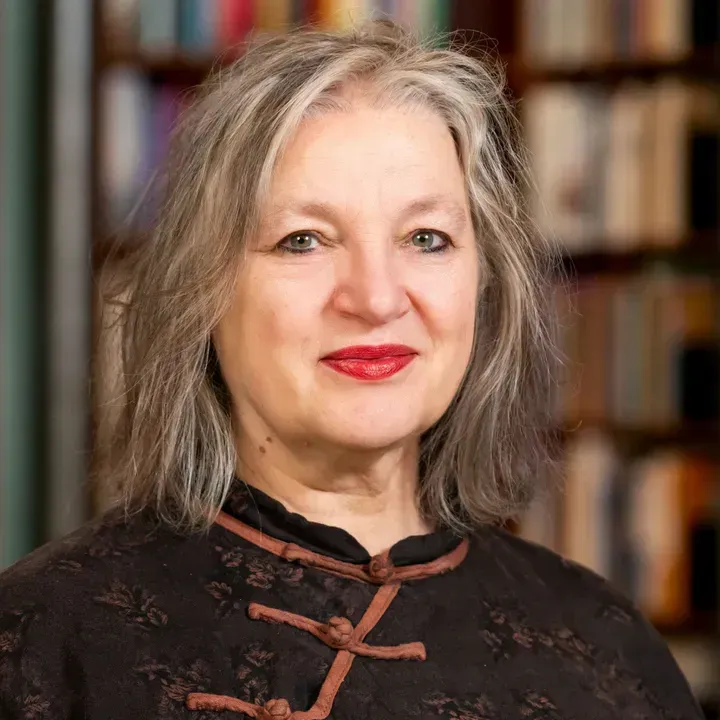 Portret van een lachende vrouw met grijs haar in een donkere kanten top, staande voor wazige boekenplanken.