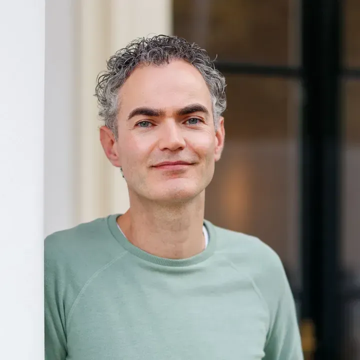 Een man in een lichtgroen shirt staat binnen en glimlacht vriendelijk bij een deuropening.