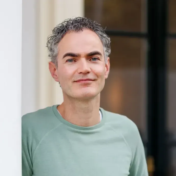 Een man in een lichtgroen shirt staat binnen en glimlacht vriendelijk bij een deuropening.