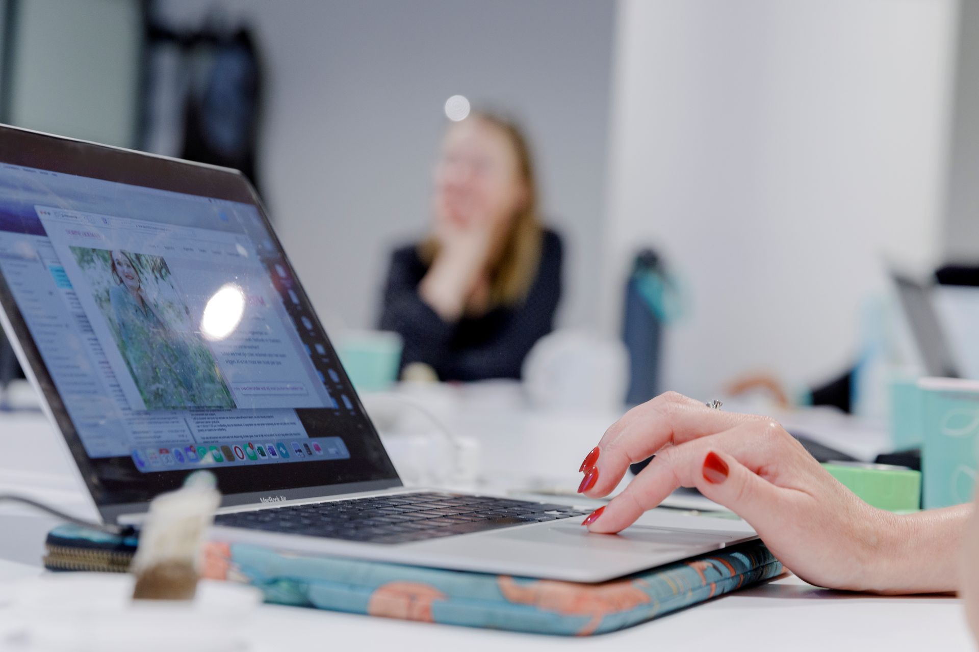 Een vrouw met rode nagels gebruikt een laptop op een tafel in een kantooromgeving; een wazig persoon op de achtergrond.