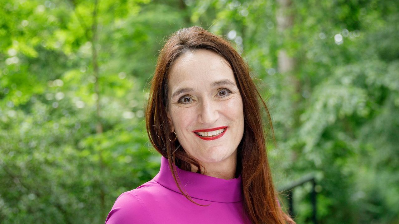 Een vrouw met lang bruin haar lacht voor het groene gebladerte, gekleed in een roze coltrui.