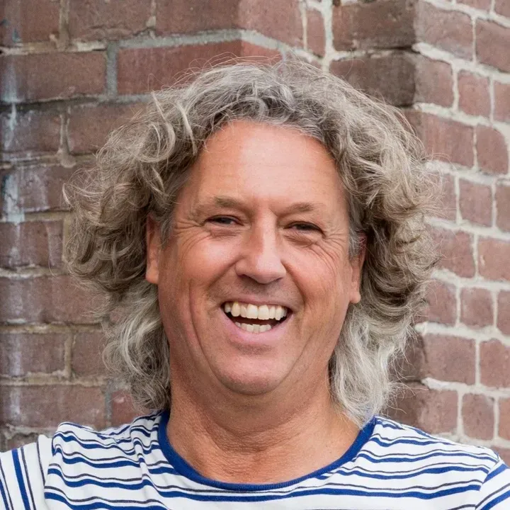 Een lachend persoon met krullend grijs haar in een gestreept shirt tegen een bakstenen muur.