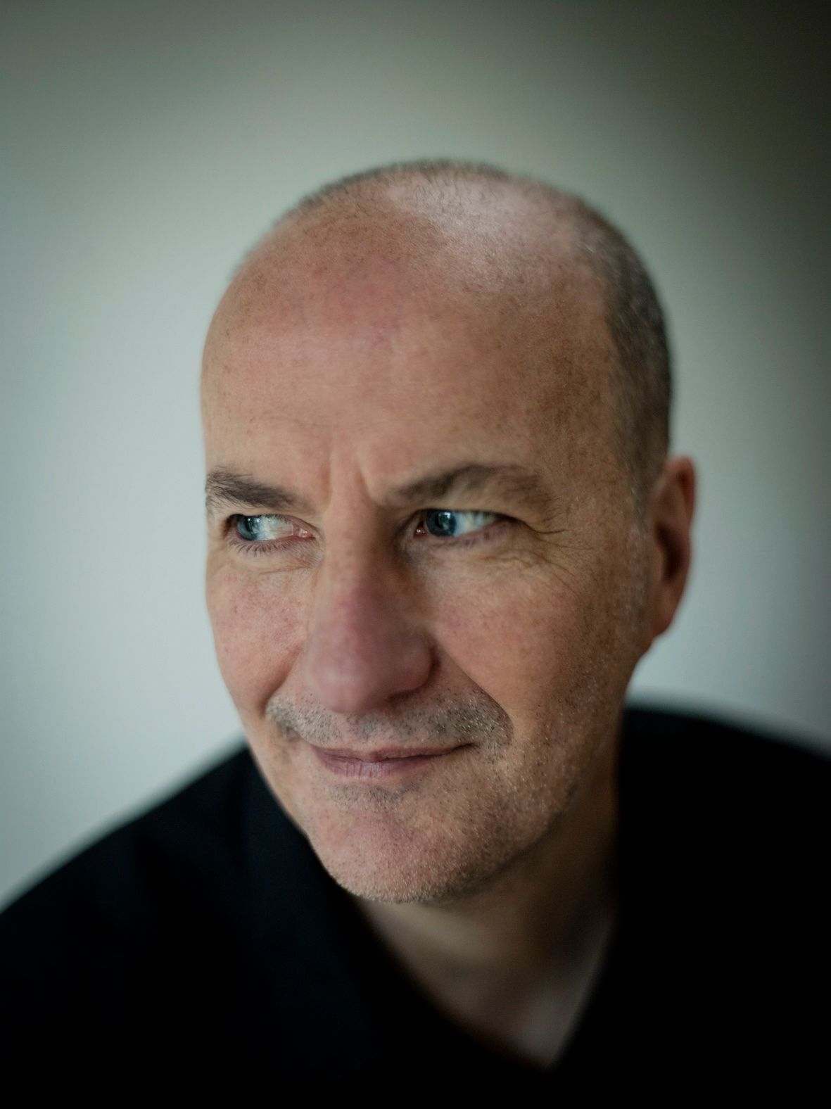 Man met geschoren haar, kijkt weg van de camera, van dichtbij gefotografeerd, met zwart overhemd aan. Gezicht scherp, achtergrond wazig.