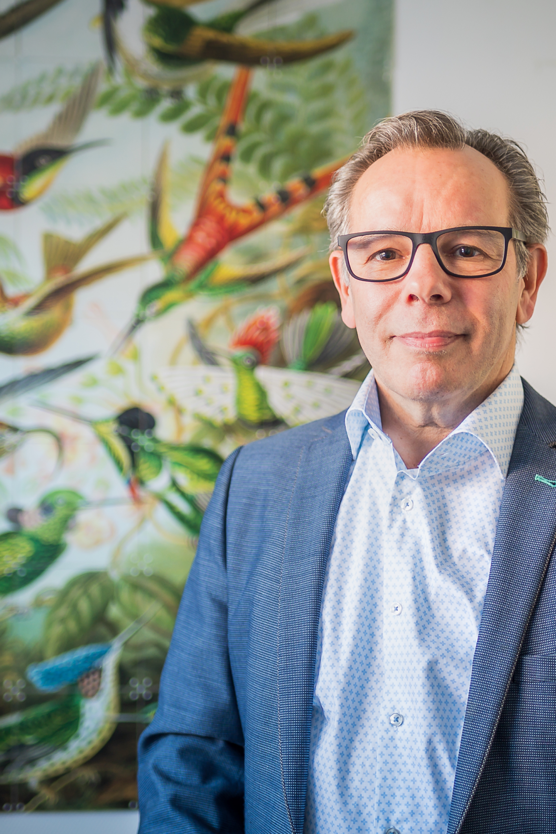 Een man in pak en bril staat voor een schilderij van vogels.