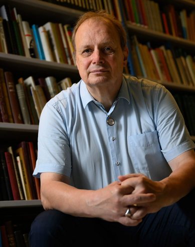 Een man zit voor een boekenkast, gekleed in een lichtblauw shirt, met zijn handen ineengevouwen.