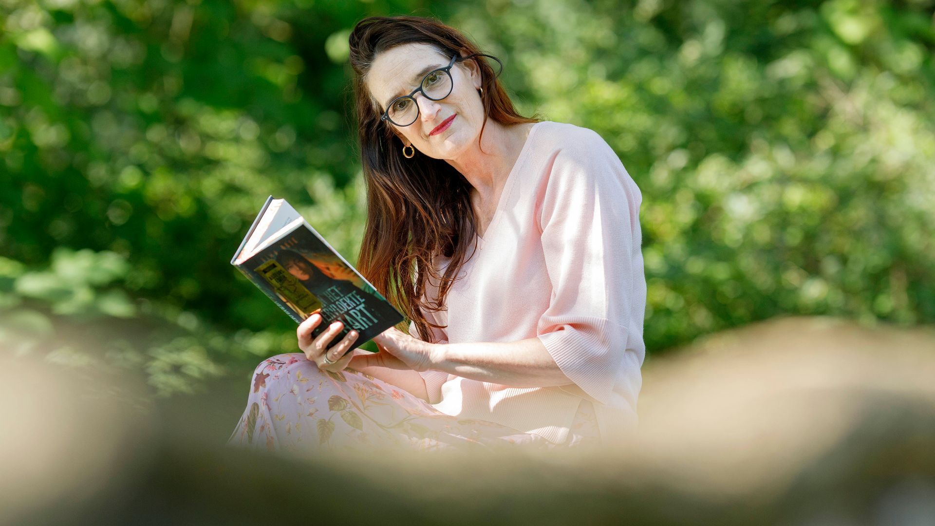 Vrouw met bril leest buiten een boek, gekleed in een roze gewaad, zonnige dag.