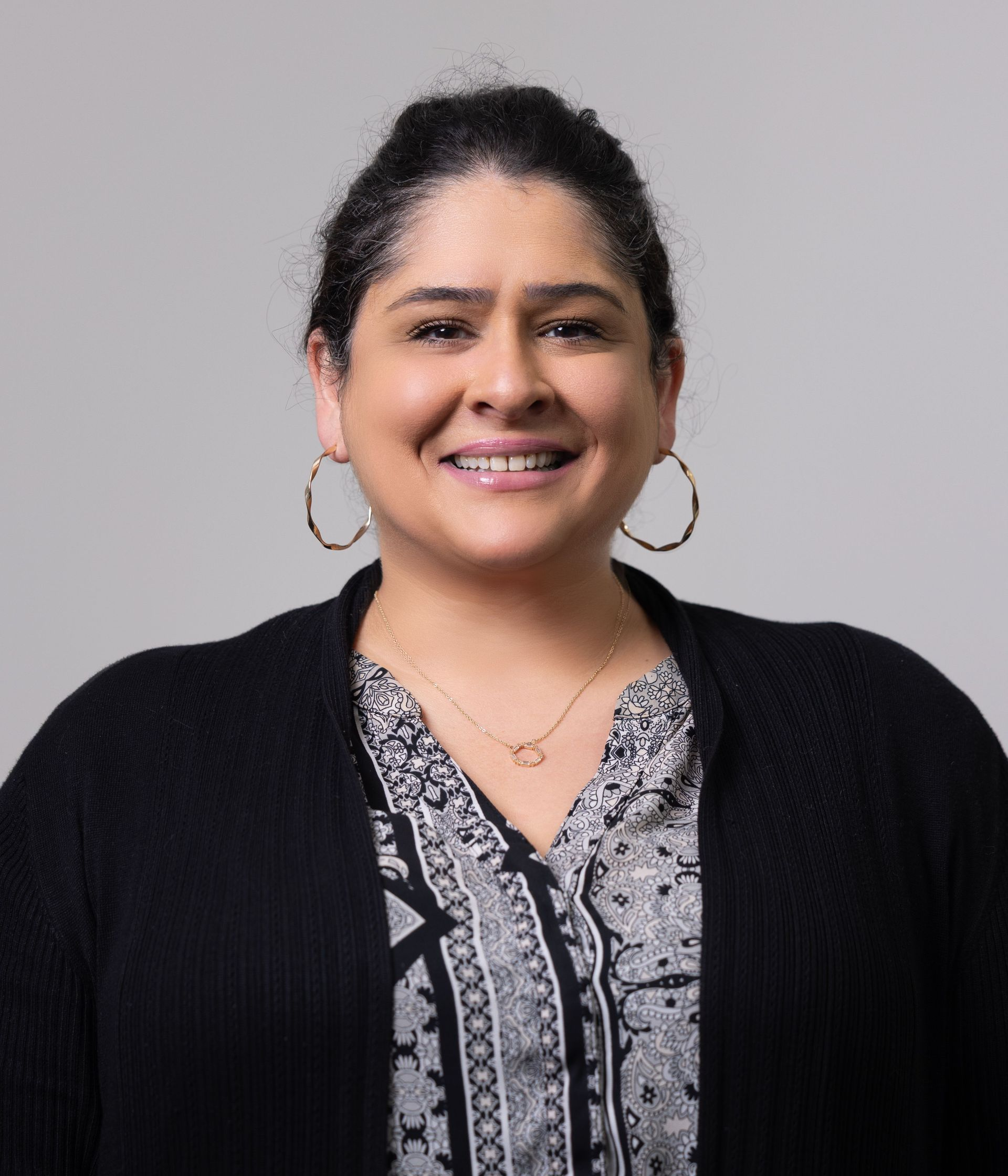 Woman smiles, wearing hoop earrings, patterned shirt, black cardigan, and gold necklace.