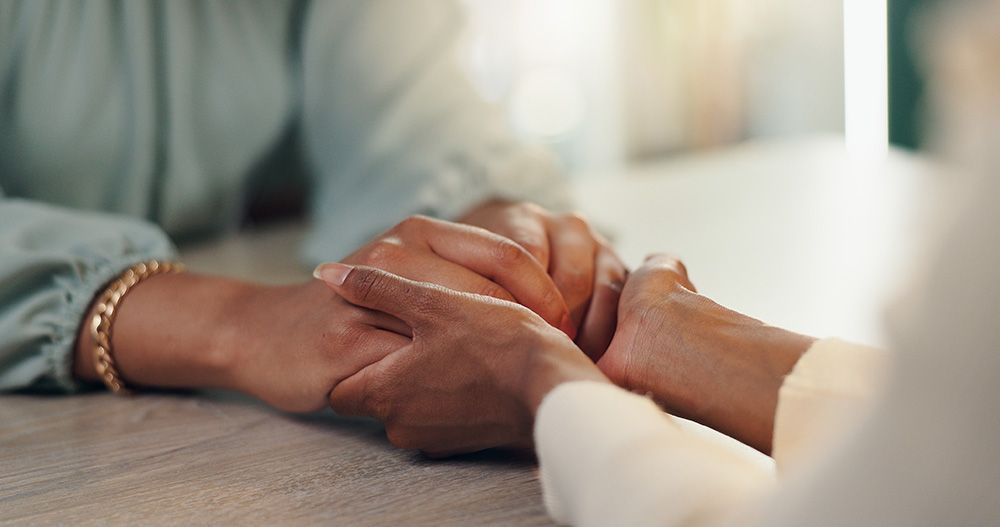 Hands of two people clasped together on a table, offering support and comfort.