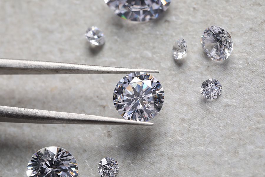 Tweezers holding a sparkling round diamond, surrounded by other loose diamonds on a light gray surface.