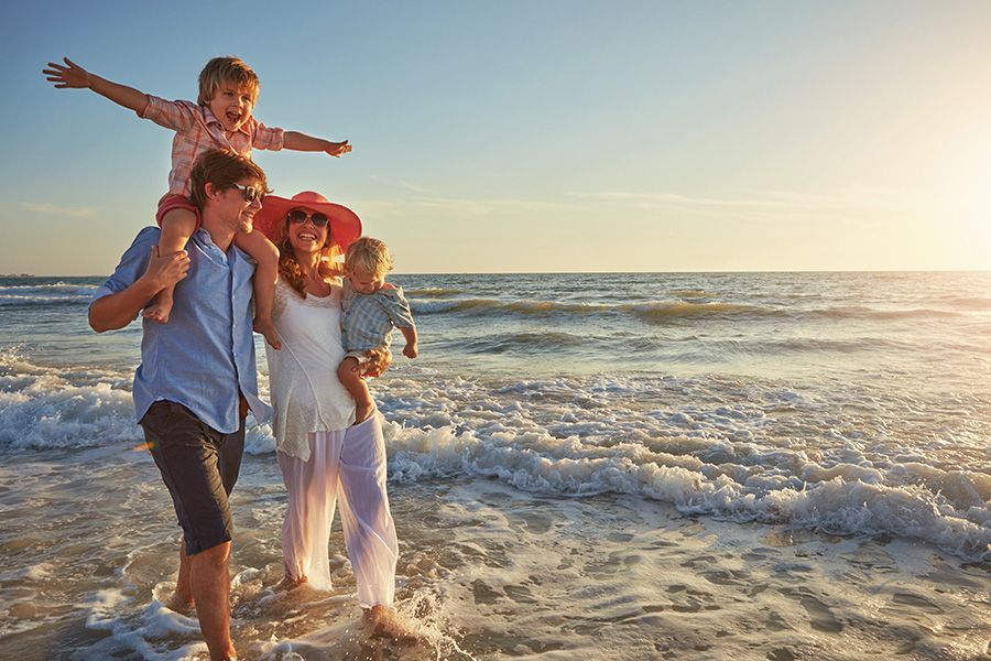 Family walking on a beach at sunset, father carrying a child on his shoulders, mother holding a baby.