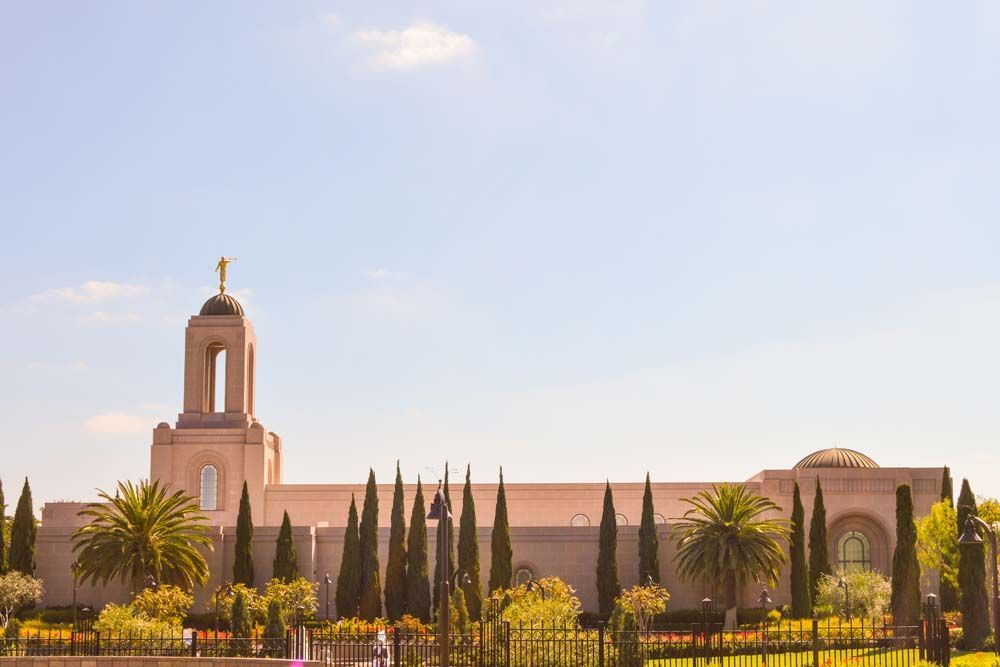 Beige building with a tall steeple, gold statue, and tall, dark green trees under a clear, blue sky.