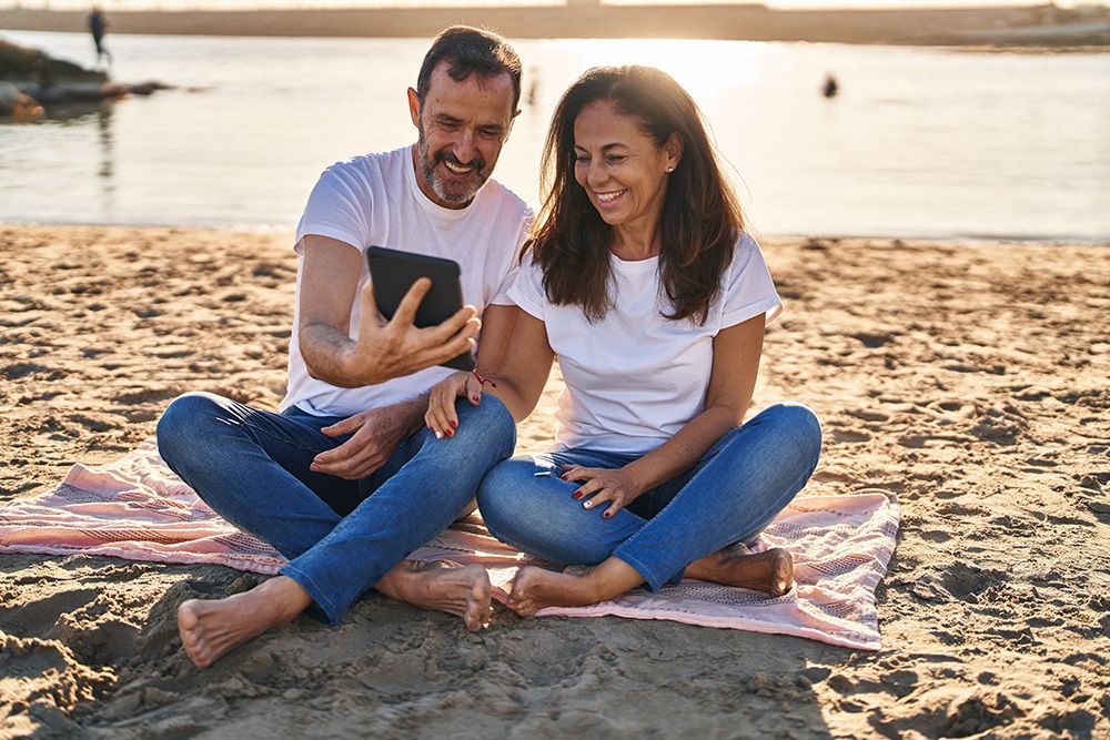 Couple sitting cross-legged on a beach blanket, looking at a tablet, smiling.