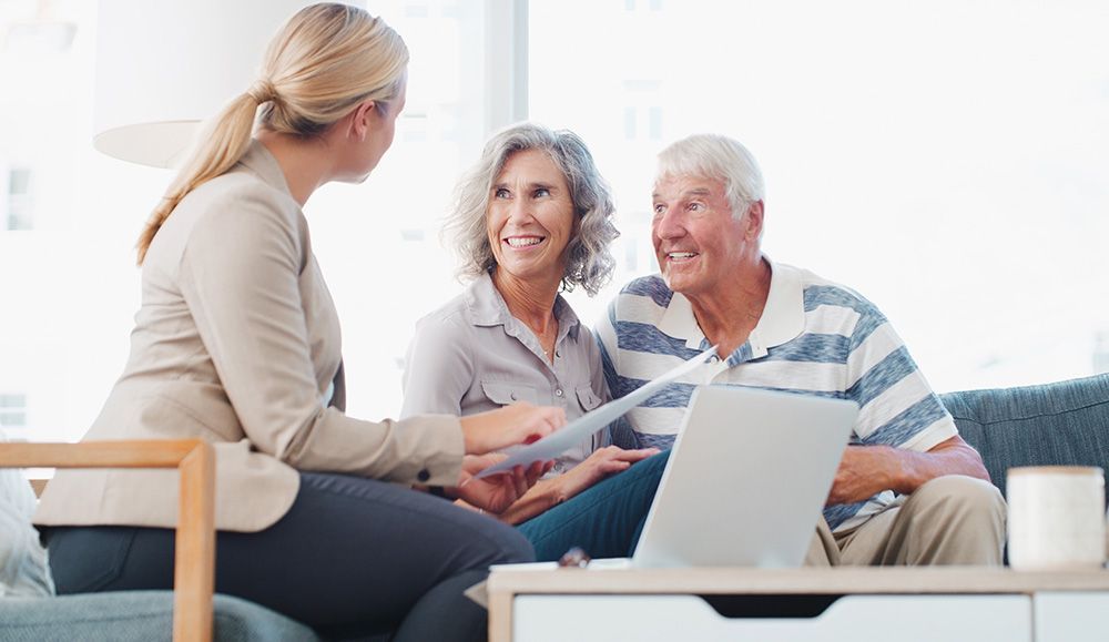 Woman showing documents to senior couple on a sofa; laptop on table.
