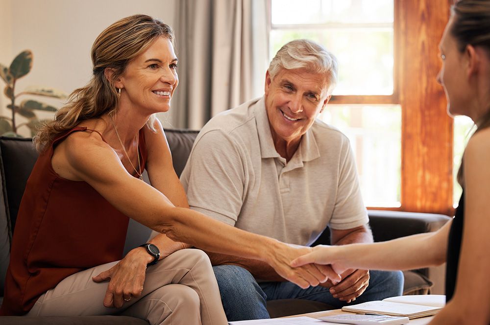 Couple shaking hands with a person, smiling. Indoors, seated on a couch.
