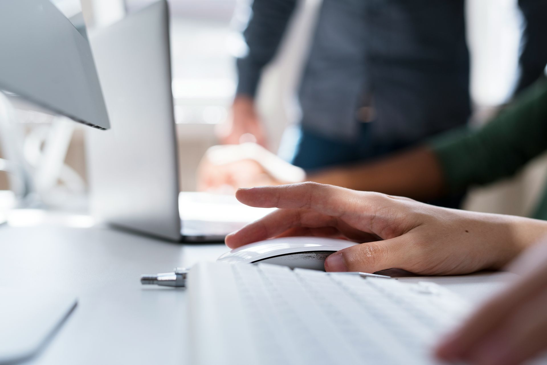 Hand clicking a mouse on a table with computers and keyboards while collaborating employees are blurred in the background.