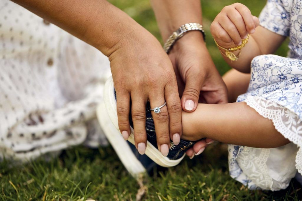Adult hand adjusting child's shoe outdoors on green grass. Child holds necklace, wearing blue dress.