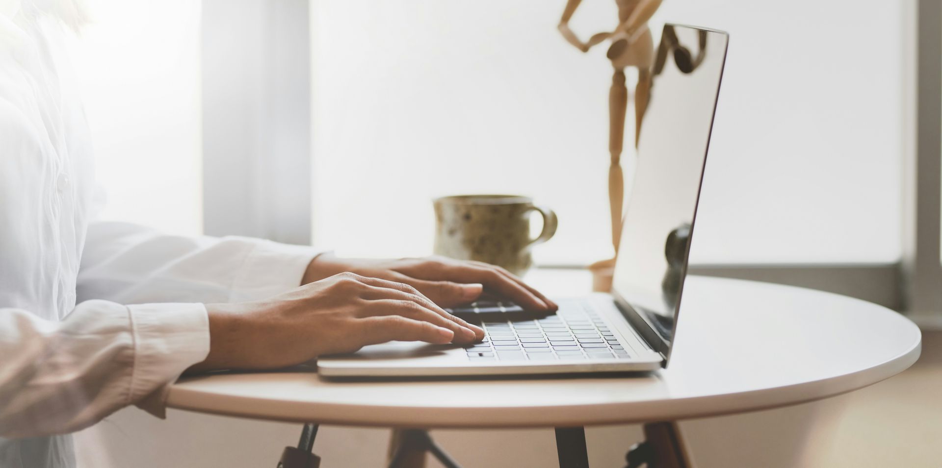 Person typing on laptop, seated at a desk near a window. Notebook and pen in the foreground.