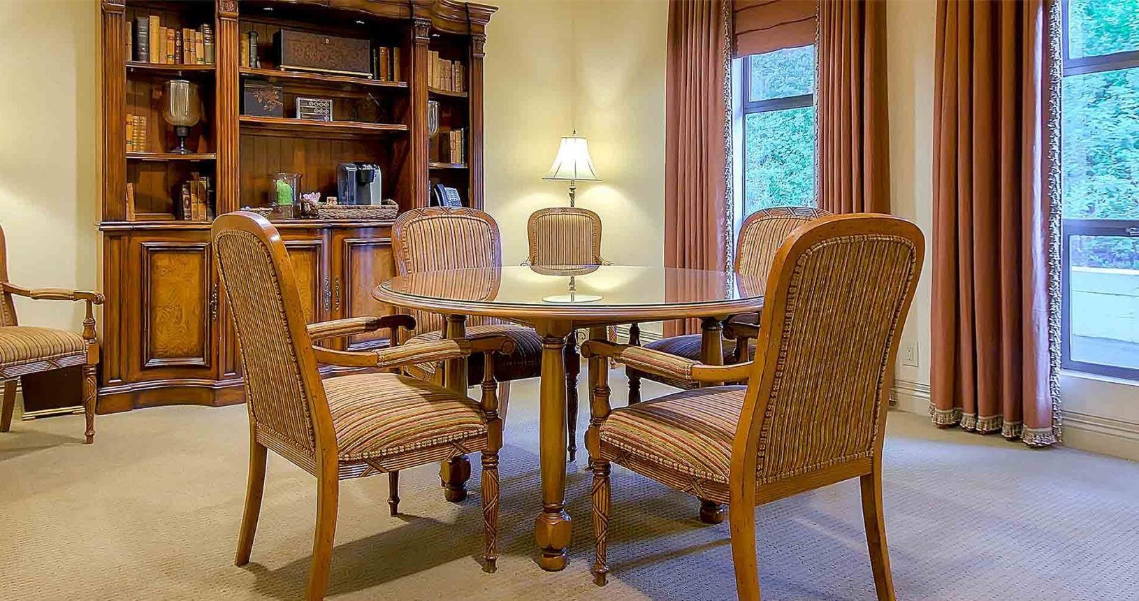 Seating area with wooden table, four wicker chairs, a bookcase, and windows with brown curtains.
