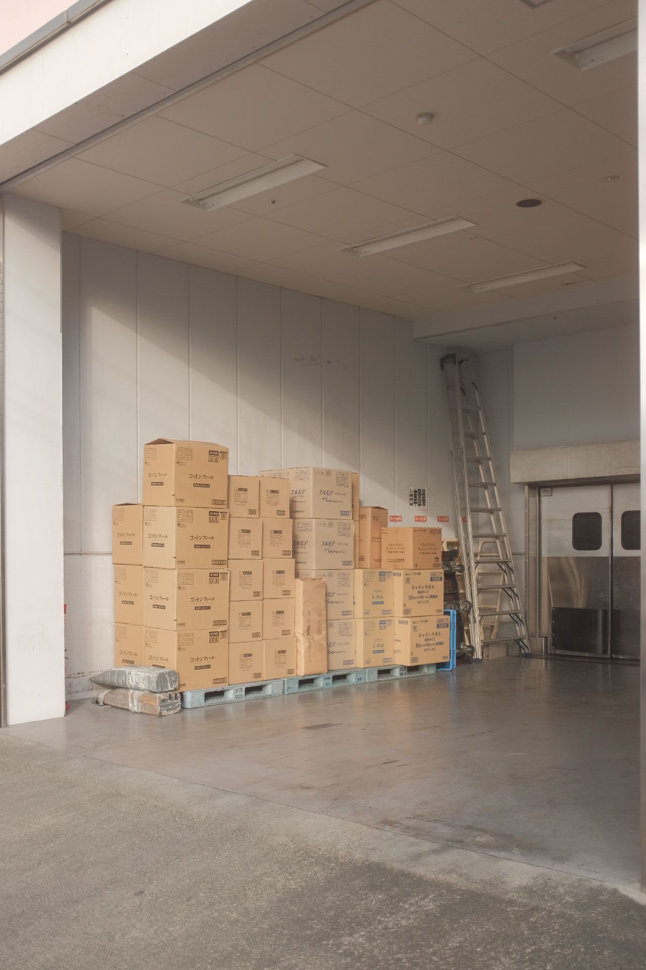 Cajas de cartón apiladas en un palé en un almacén, con una escalera y una puerta visibles.