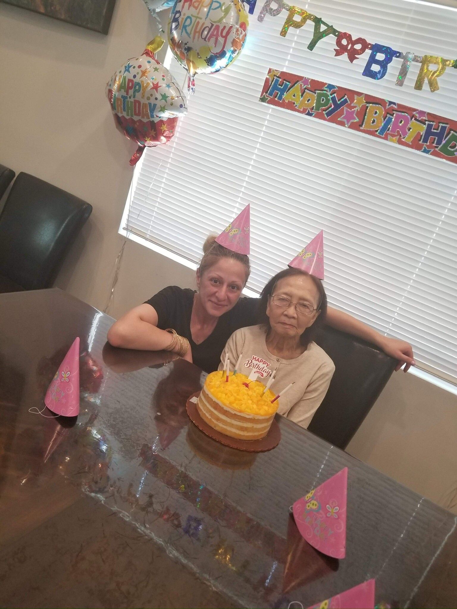 Two women are sitting at a table with a birthday cake and balloons.