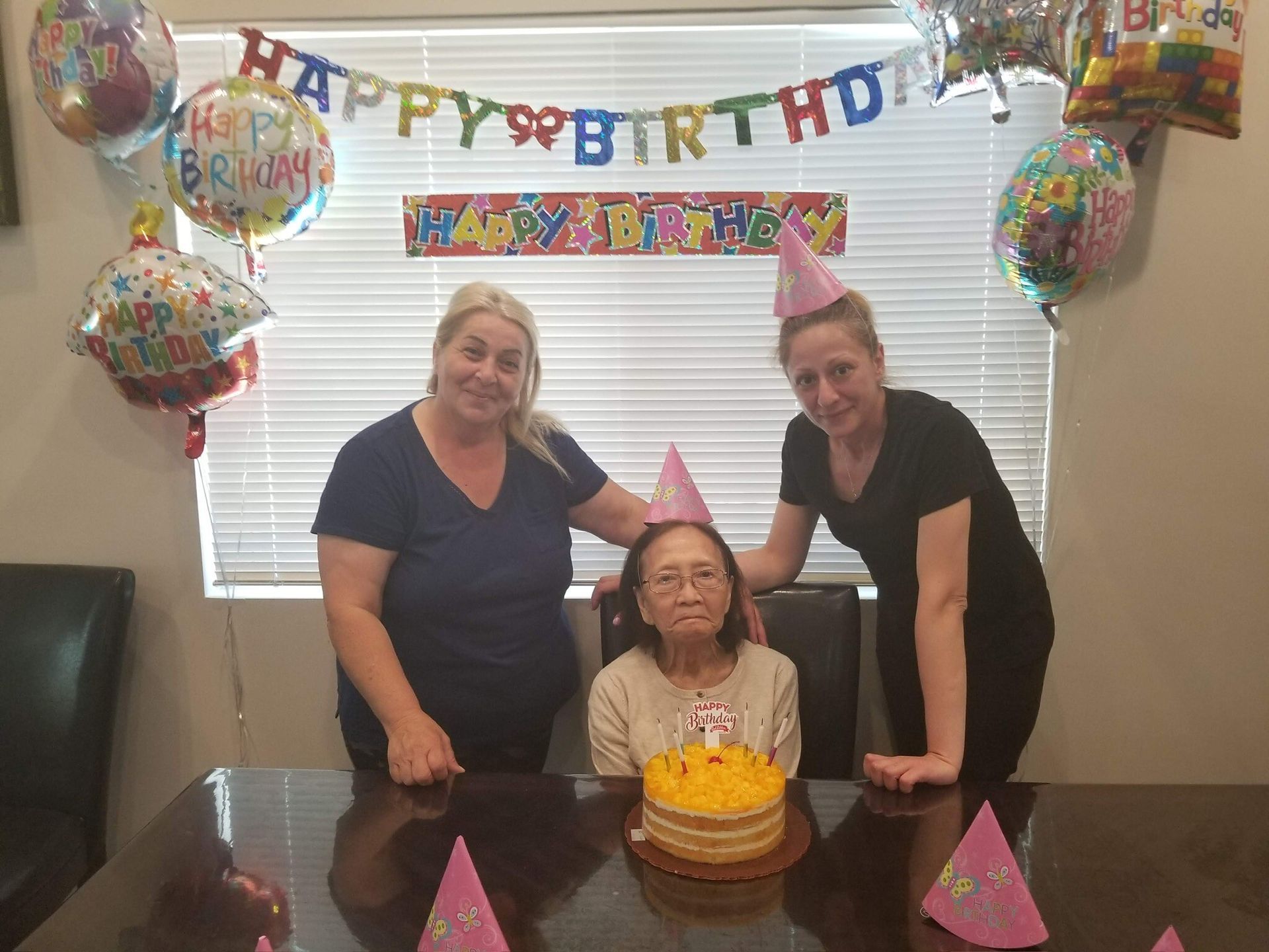 Three women are sitting at a table with a birthday cake and balloons.