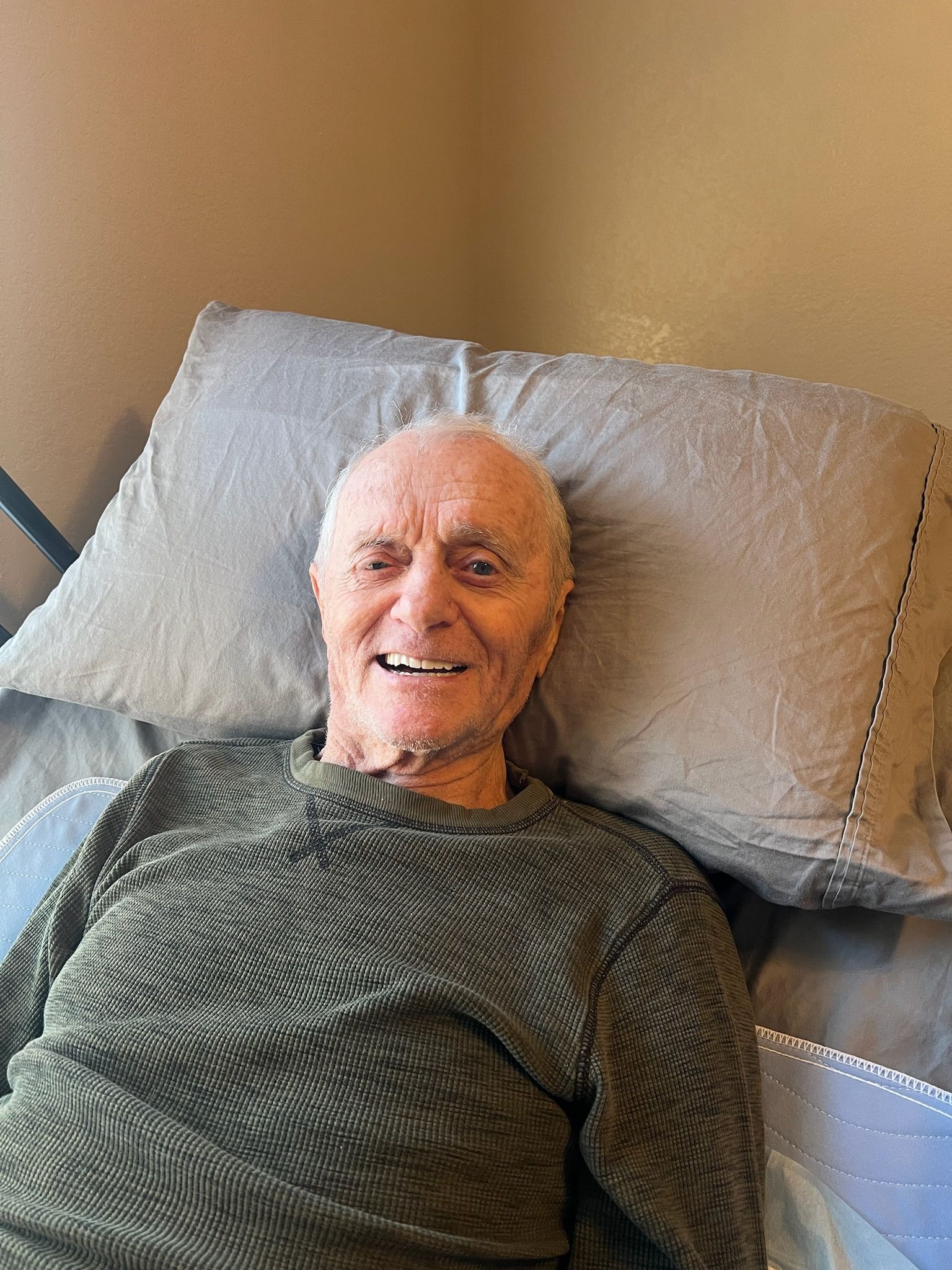 An elderly man is laying in a bed with a pillow and smiling.