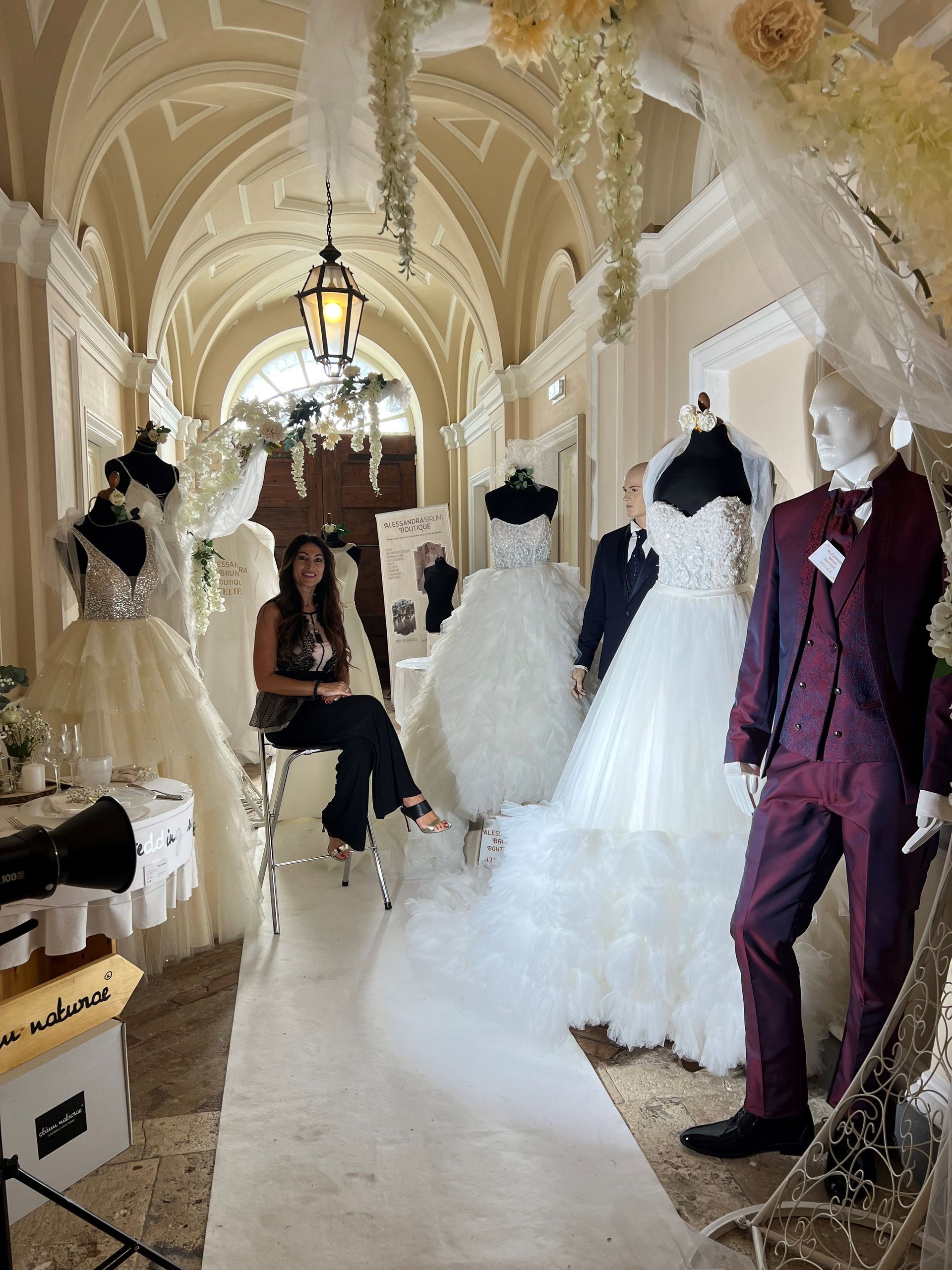 Una persona siede in un corridoio pieno di abiti da sposa su manichini e un abito formale, decorato con tessuto bianco.