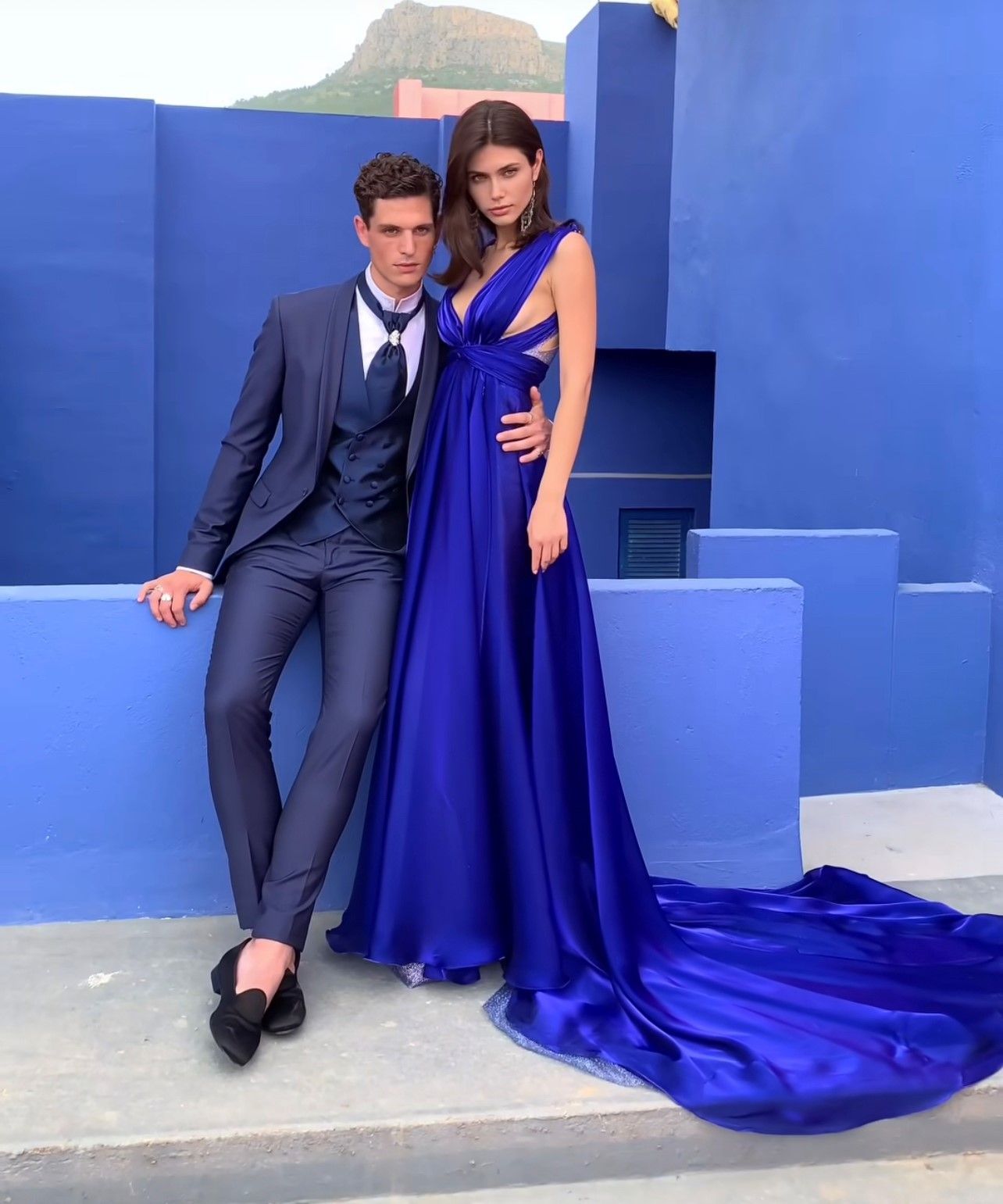 In un ambiente dai colori vivaci, un uomo in abito blu scuro si appoggia a una parete blu accanto a una donna che indossa un lungo e fluente abito blu reale.