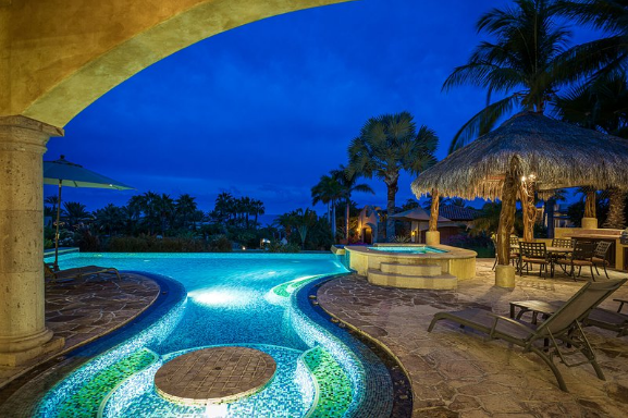 A large swimming pool is lit up at night with a gazebo in the background.