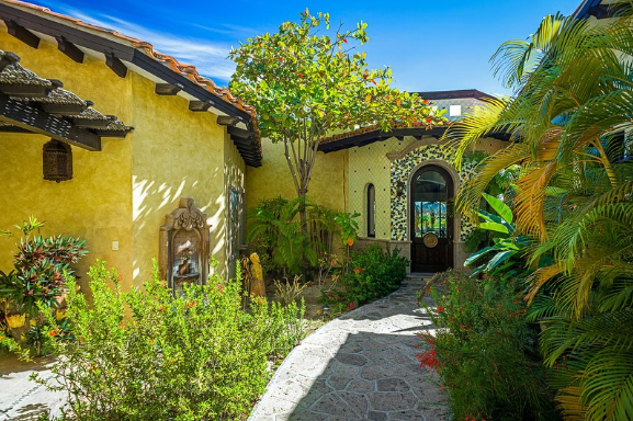 A yellow house with a stone walkway leading to it surrounded by trees and bushes.