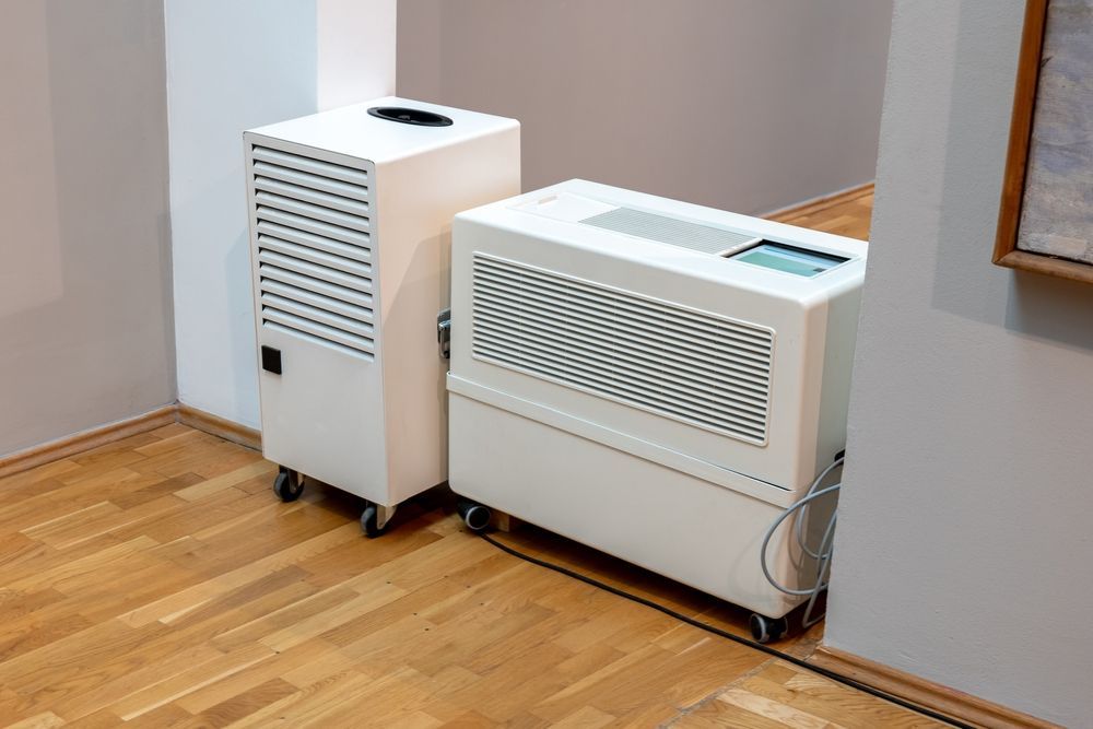 Two white air conditioners are sitting next to each other on a wooden floor in a room.