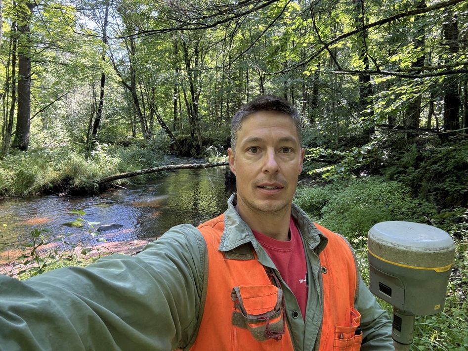Person taking a selfie in a forest beside a stream, wearing an orange safety vest