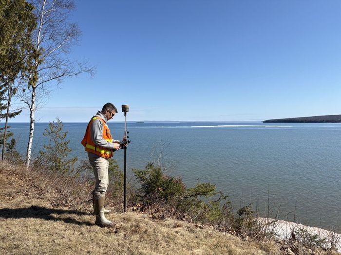 Person in an orange safety vest using a surveying tool beside a lake on a sunny day