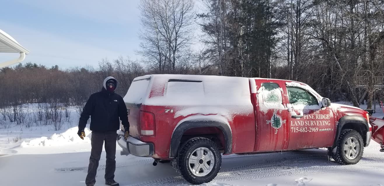 A person in a black hoodie stands in a snow-covered yard next to a red pickup truck with a white camper shell.