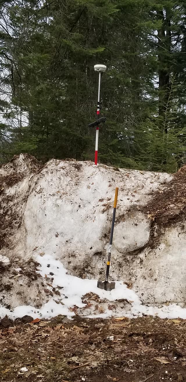 A survey rod with a GPS receiver stands atop a snow pile next to a shovel in a wooded area.