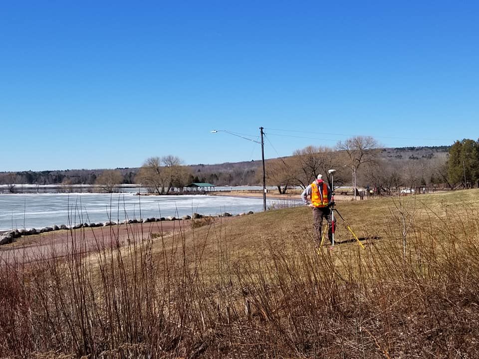 A person in a high-visibility vest uses surveying equipment on a grassy hill beside a frozen lake on a clear day.