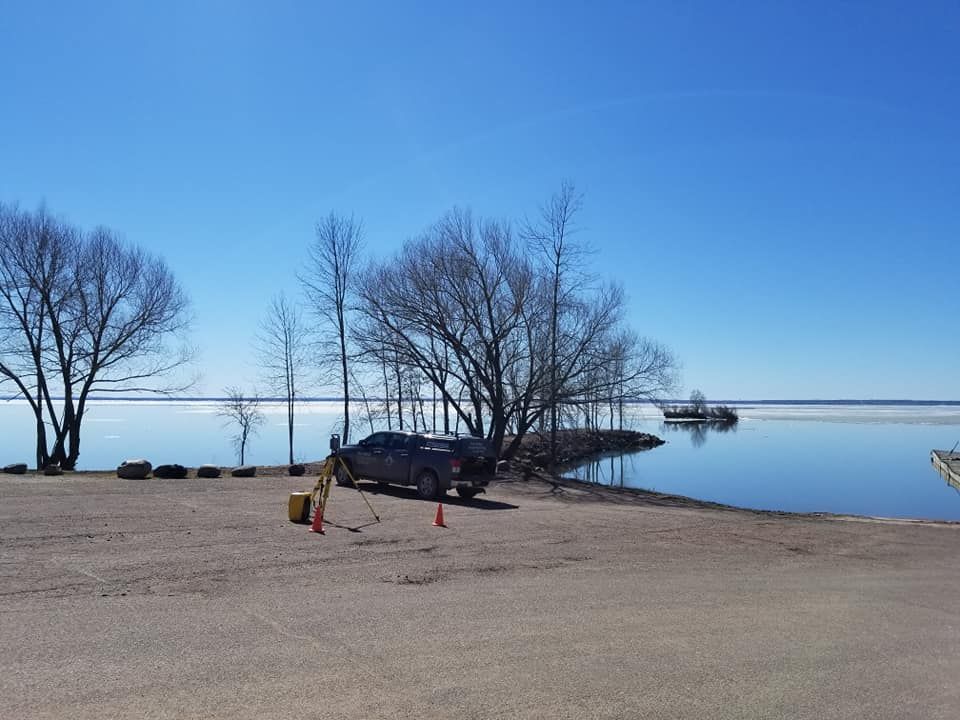A gravel parking area by a calm lake with bare trees under a clear blue sky, featuring a pickup truck and two orange cones.