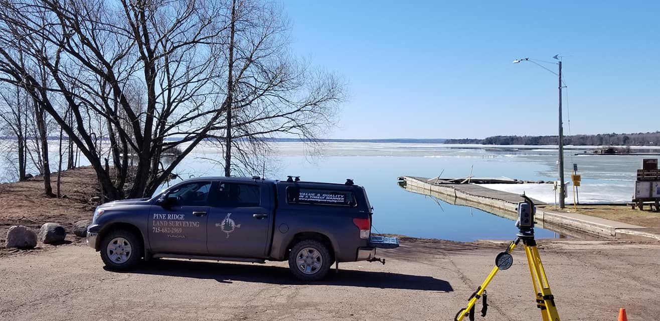 A grey pickup truck parked by a lake on a sunny day, with a surveying tripod set up in the foreground.