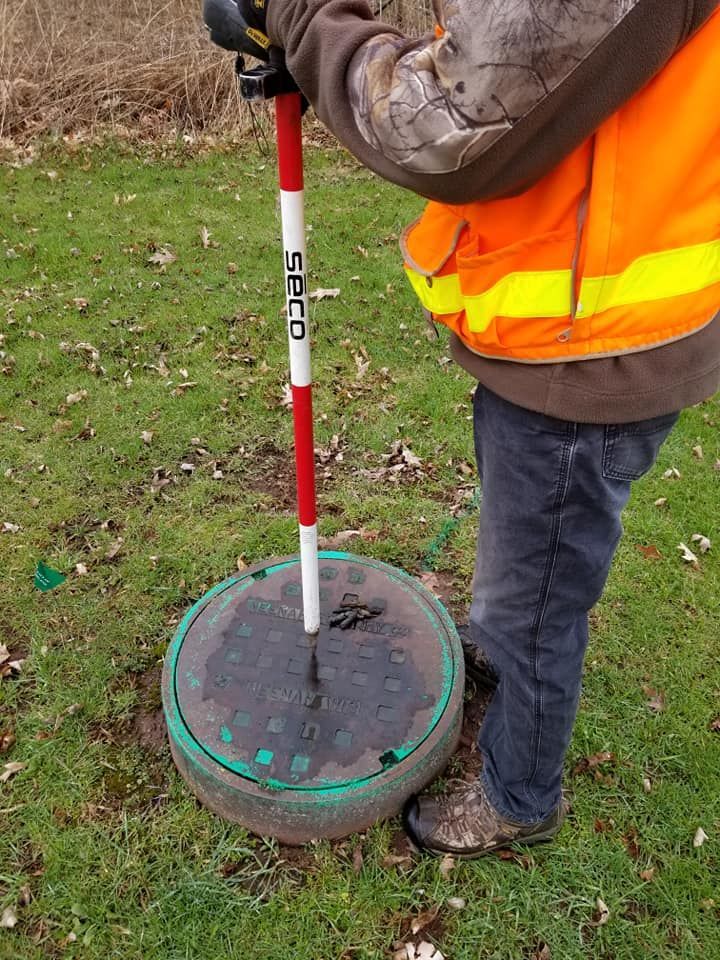 A person in a high-visibility orange vest uses a surveying pole to mark the center of a circular manhole cover in a field.