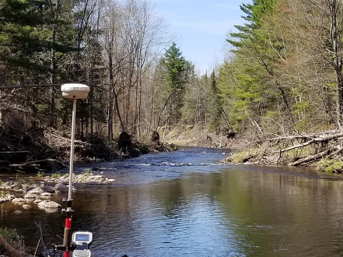 A GNSS surveying antenna mounted on a tripod at the edge of a stream flowing through a forested area on a sunny day.