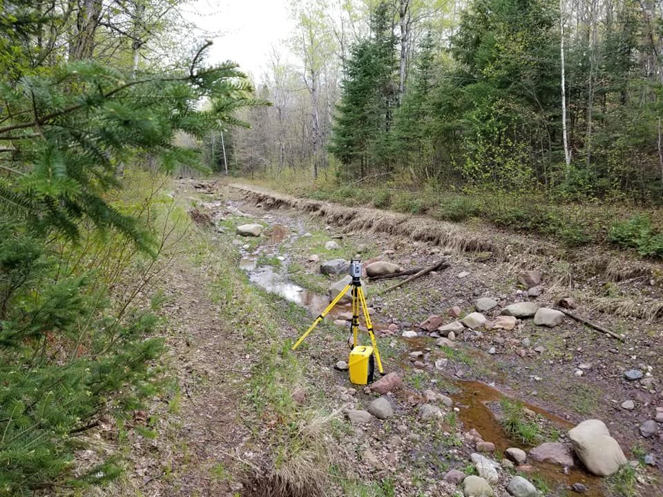 A survey instrument on a yellow tripod stands in a rocky, shallow streambed surrounded by trees and vegetation.