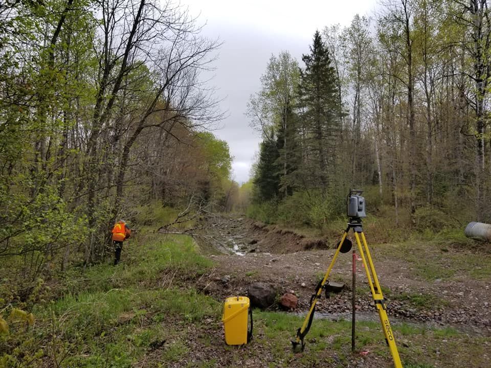 A surveyor with a bright orange backpack stands near a tripod-mounted surveying instrument in a wooded, rocky area.