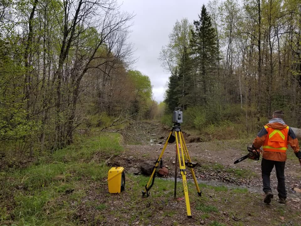 A surveyor in a high-visibility orange vest walks near a surveying instrument on a tripod in a wooded area.