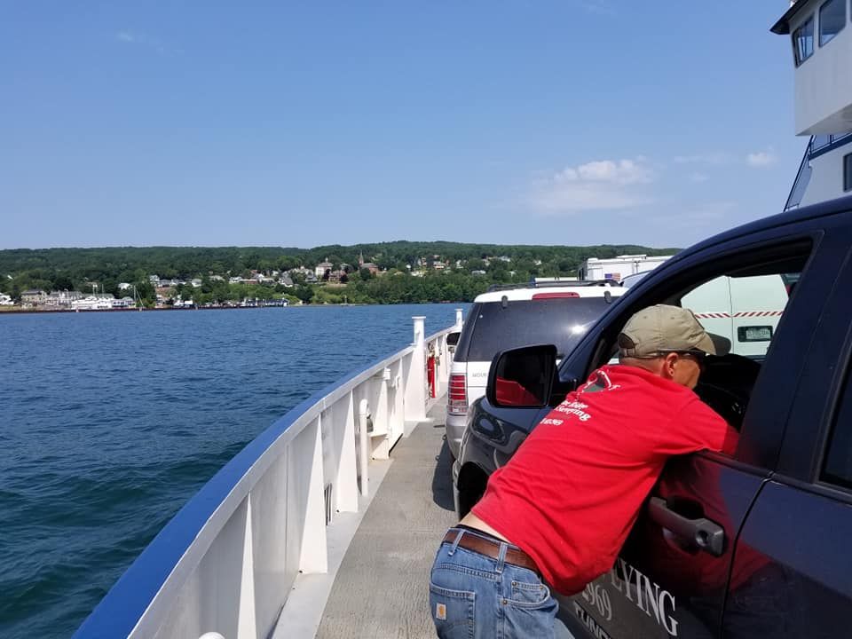 A person in a red shirt leans into the window of a car on a ferry deck with a view of a shoreline under a clear blue sky.