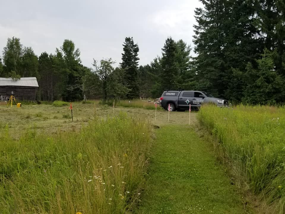 A gray pickup truck parked in a grassy field near a mowed path, with a rustic building and trees in the background.