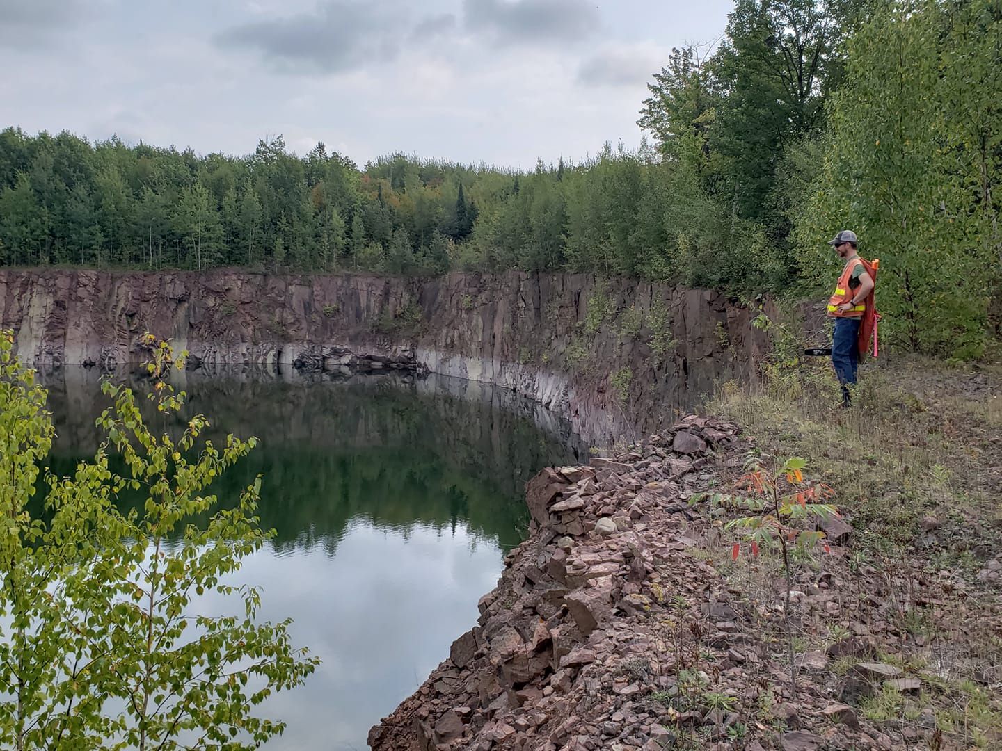 A person in a high-visibility vest stands on the rocky edge of a deep, water-filled quarry surrounded by trees.