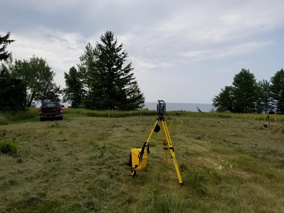 A surveying instrument on a yellow tripod stands in a mowed field near a vehicle and trees under a cloudy sky.
