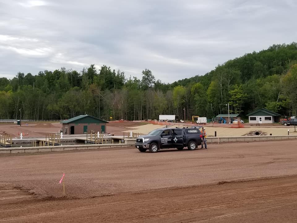 A pickup truck parked on a red dirt construction site near a small building, with a forest in the background.